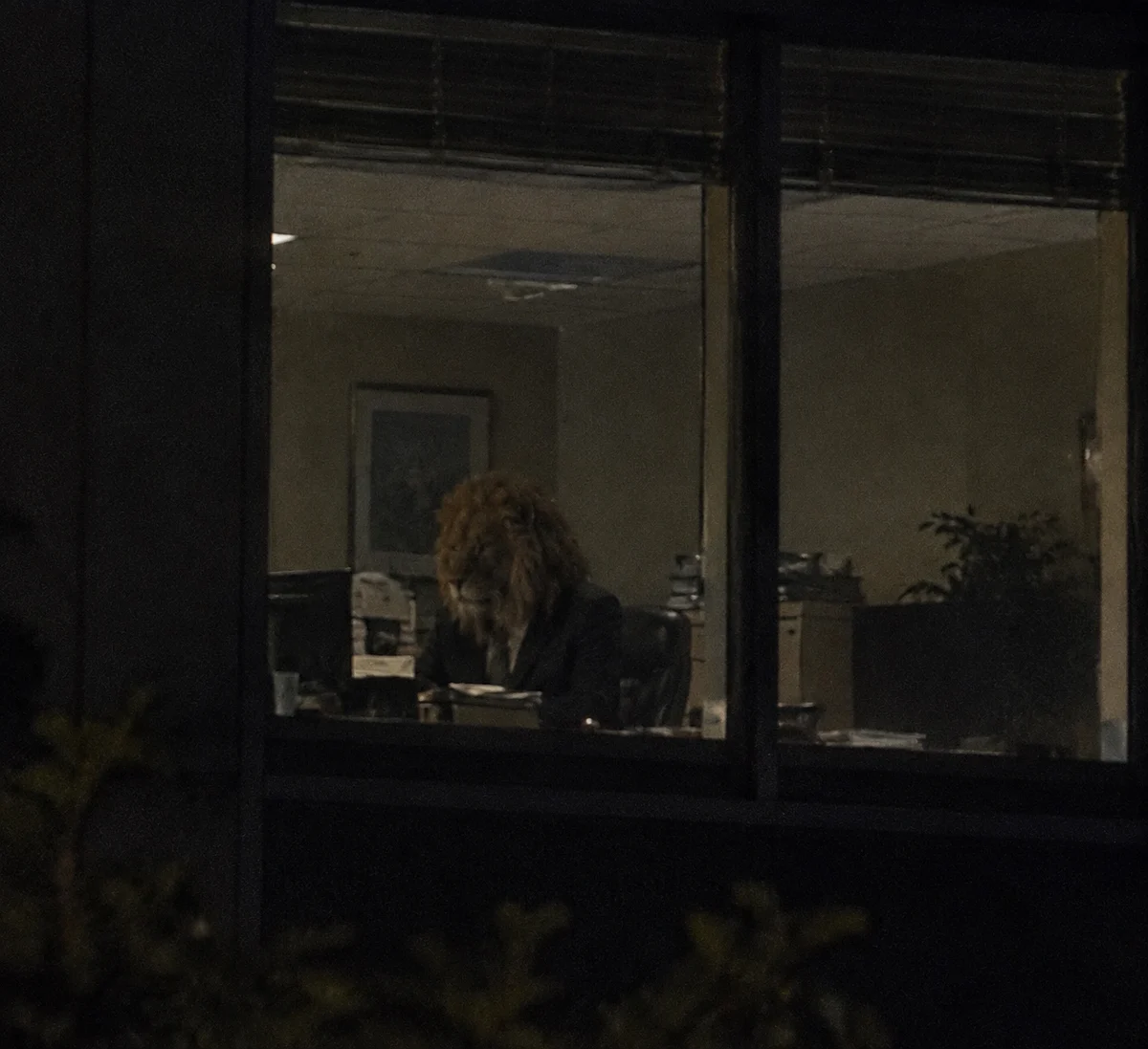 Illustration of a lion-headed figure at a desk, seen through an office window at night