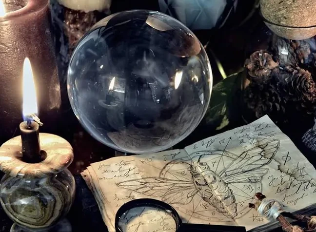 A crystal ball, lit candles, pine cones and an open spell book with a sketched winged insect on a dark altar.